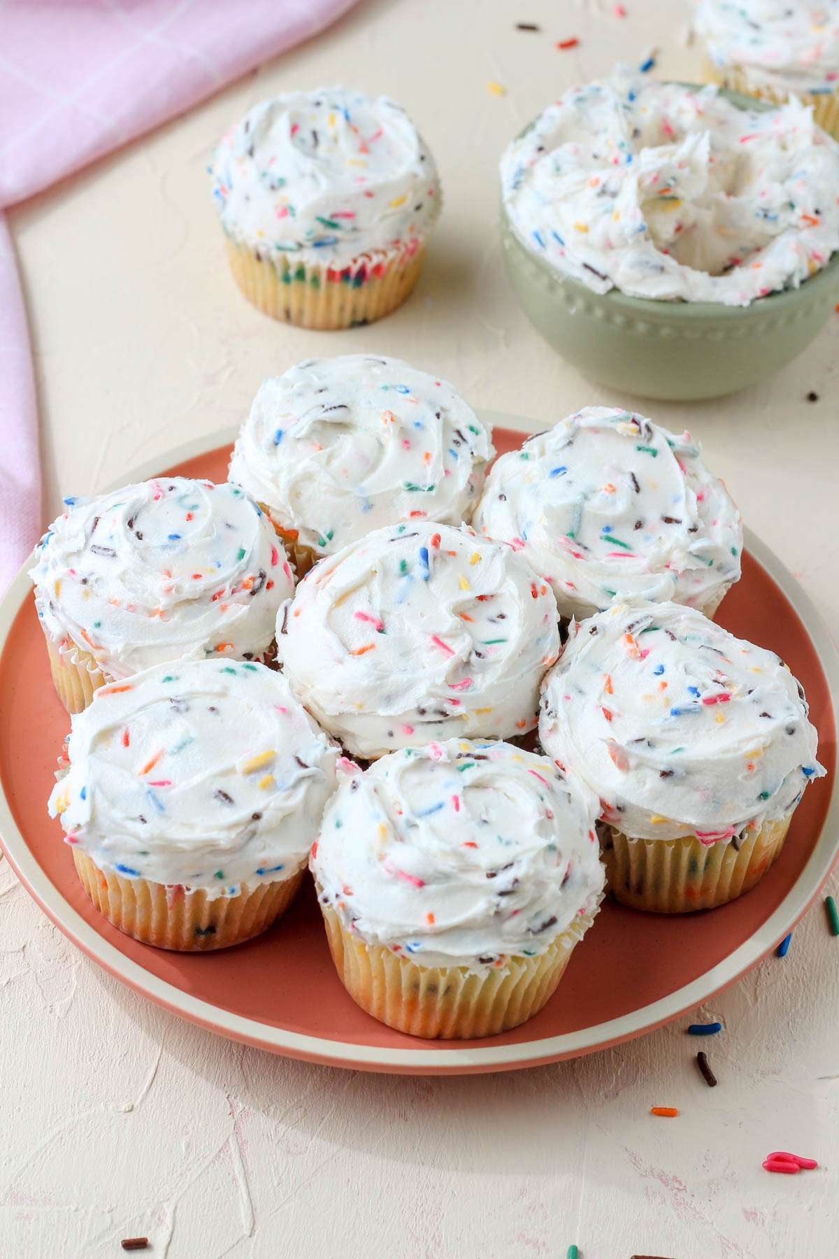 A small pink plate with iced dairy-free funfetti cupcakes on a cream counter.