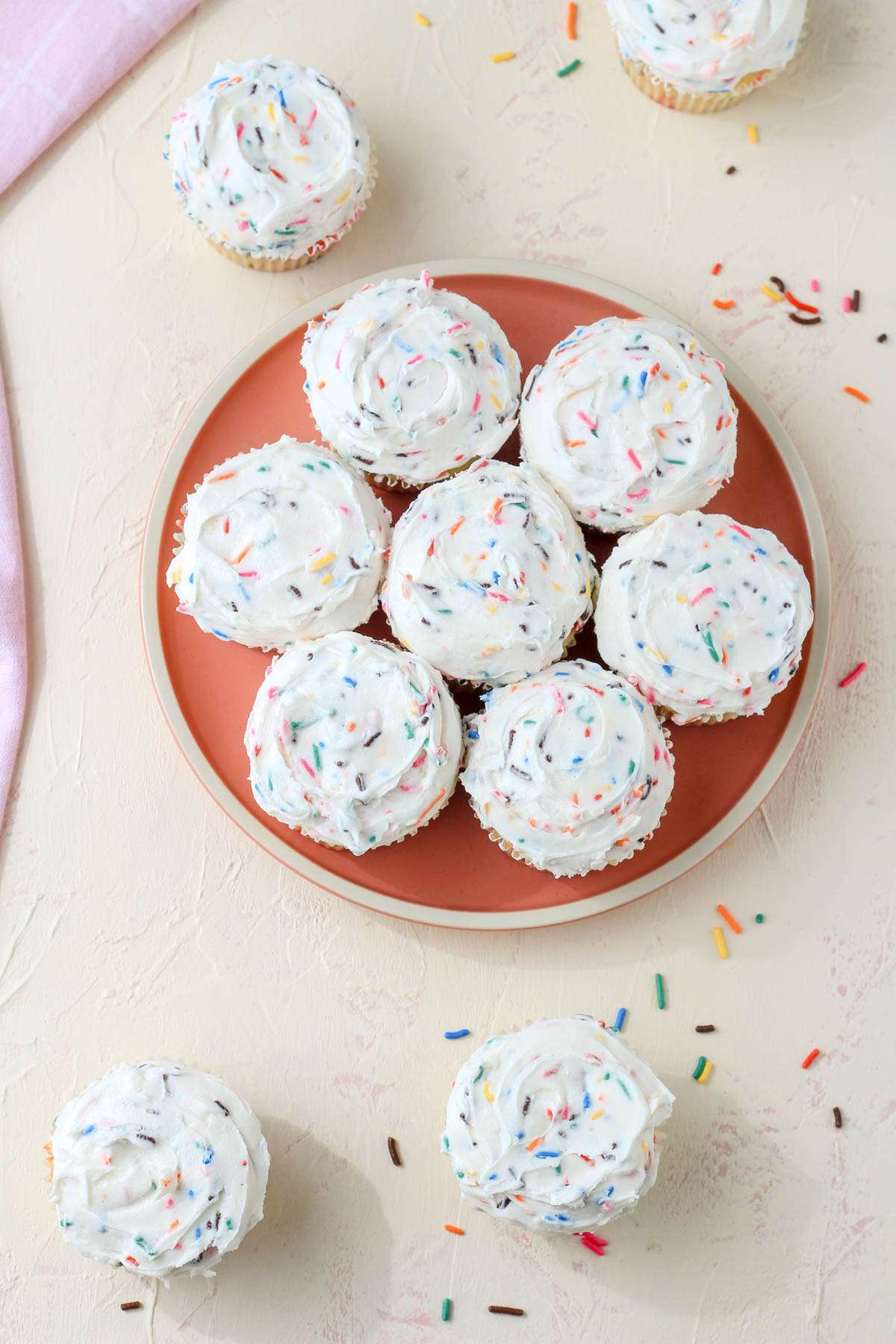 A top down image of a pink plate covered in dairy-free funfetti cupcakes iced with funfetti icing with a few cupcakes off the plate in the bottom left and top right.