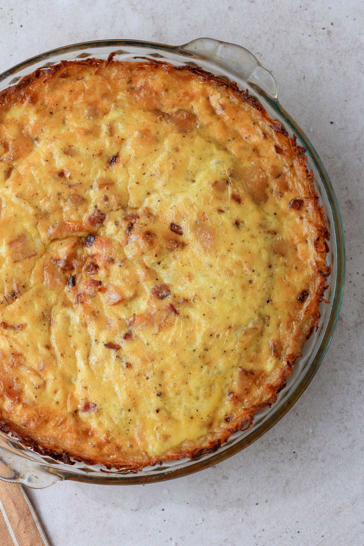 A top down of the the right 75% of a hash brown crust quiche in a glass pie dish on a tan counter.
