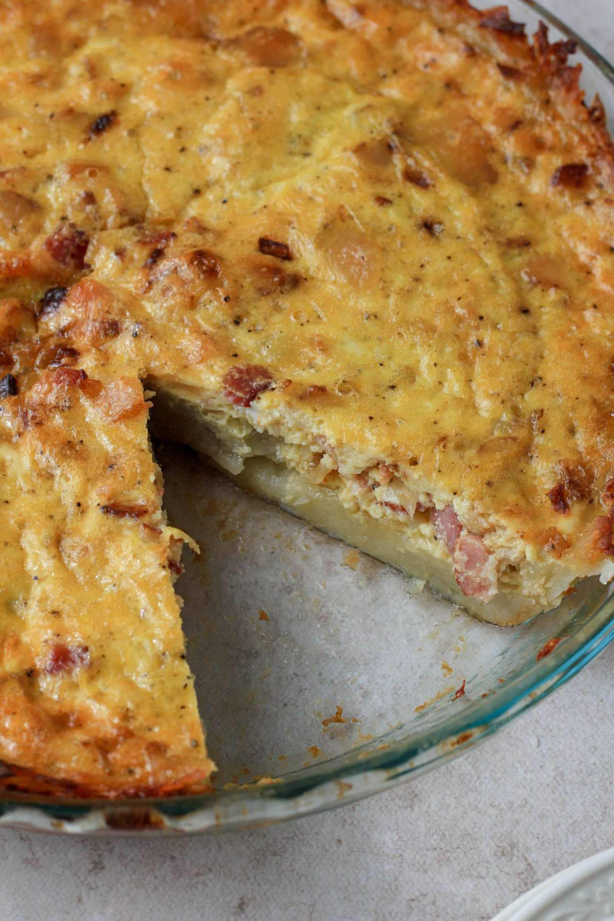 A close up of a glass pie pan with a hash brown crusted quiche after a slice has been removed.
