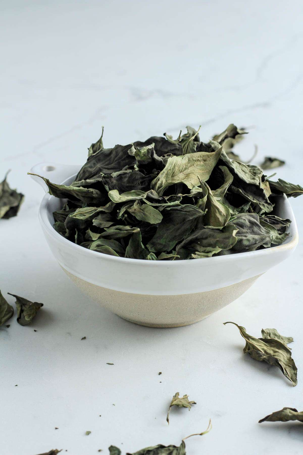 A white bowl from the side filled with dried basil leaves.