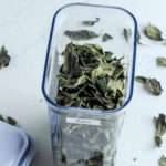 A close up of a pop top container filled with dried basil leaves on a white counter.