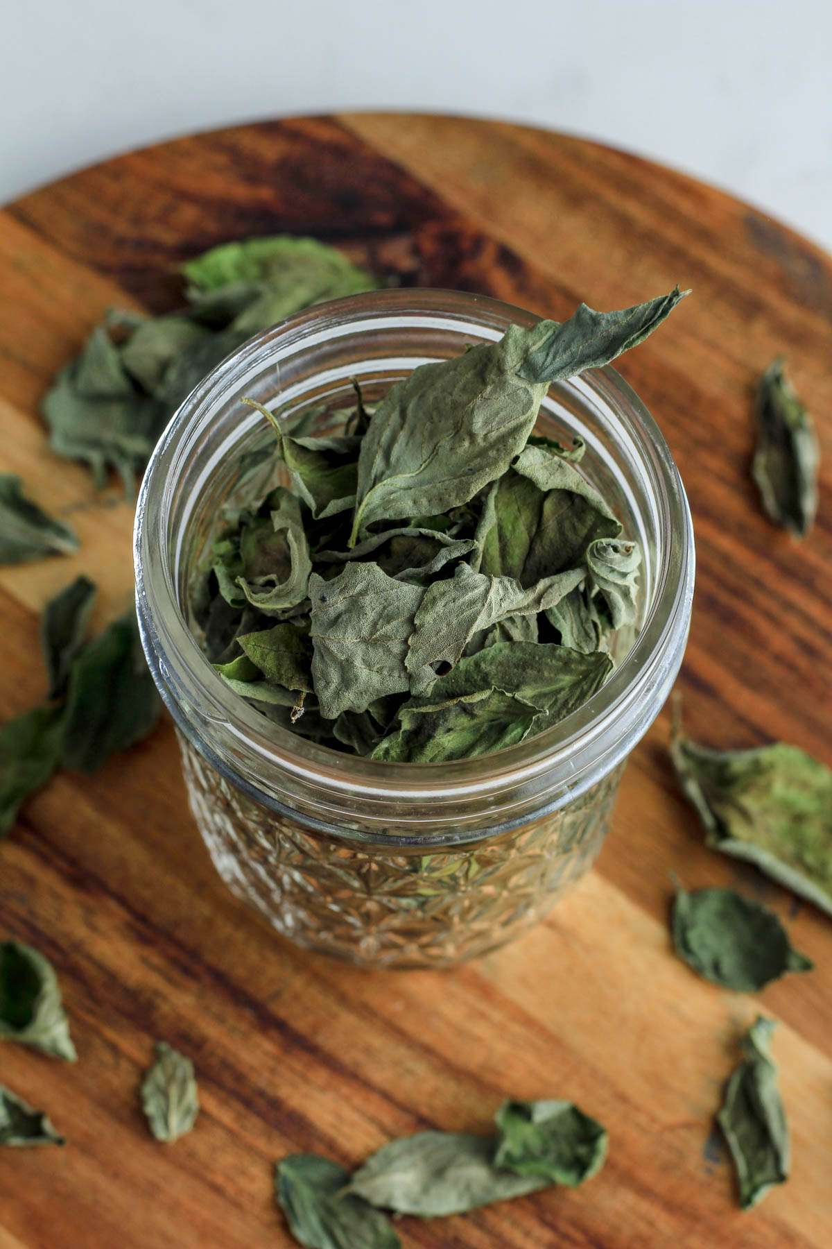 A top down image of a mason jar filled with dried basil leaves on a wooden counter with leaves scattered around the base of the jar.