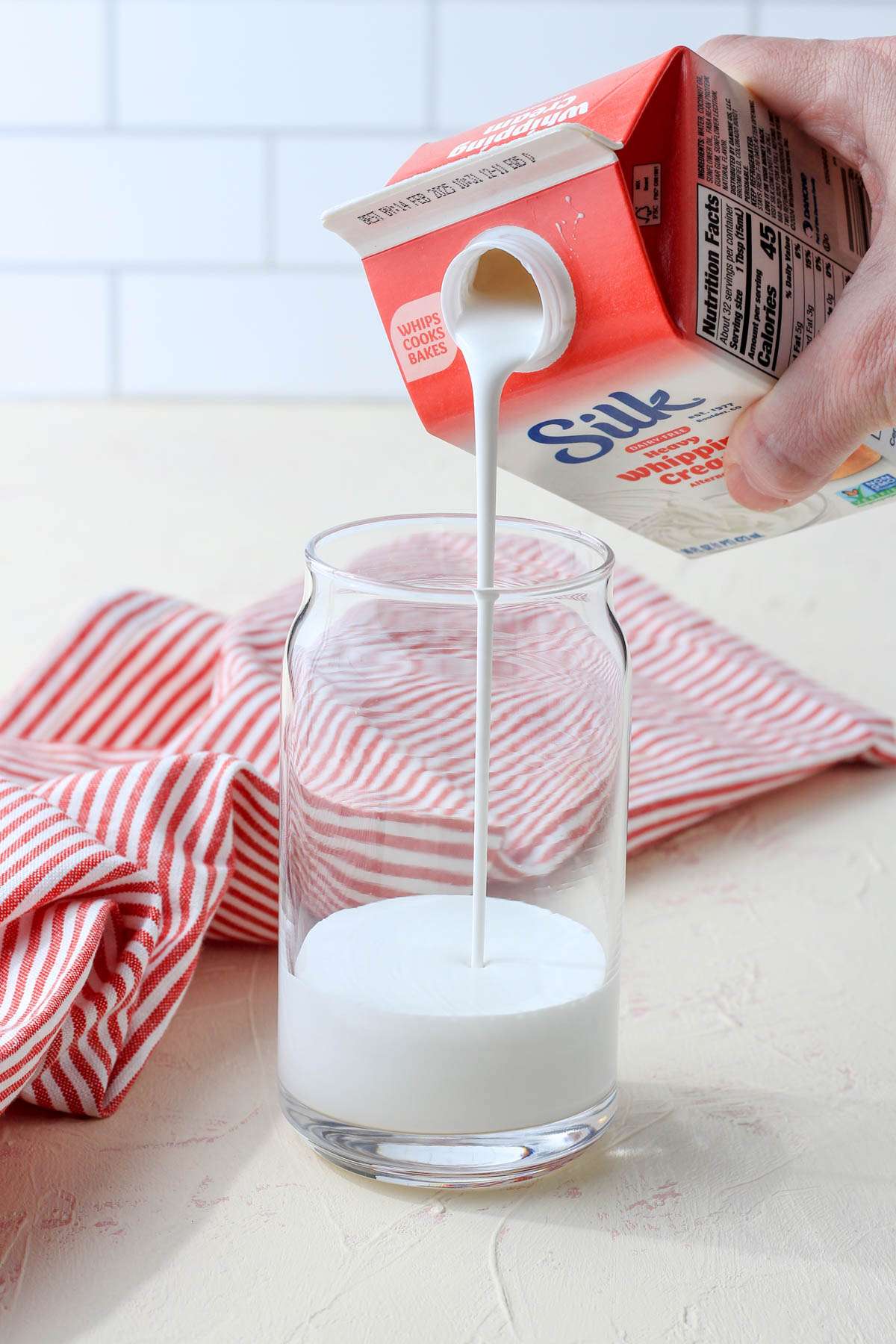 A tall glass with silk heavy whipping cream being poured out of the carton and into the glass.