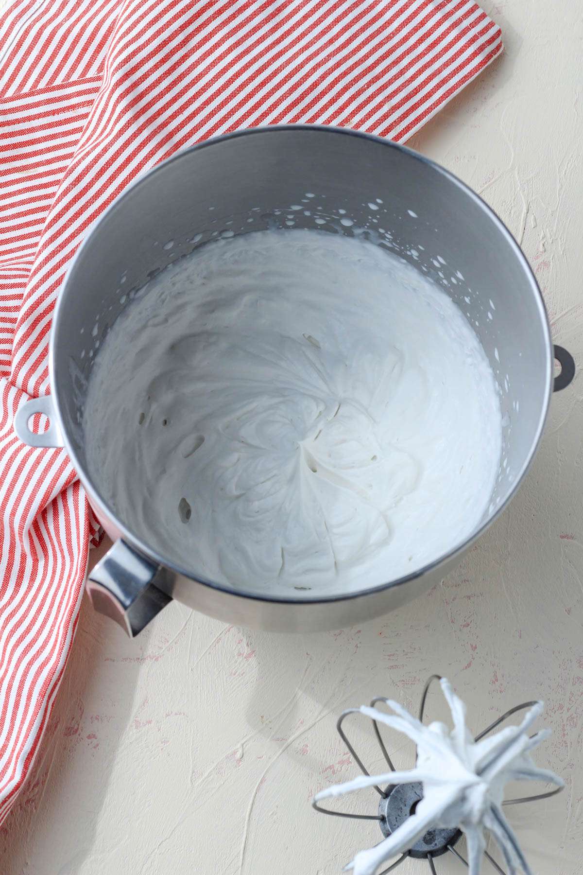 A silver mixing bowl with whipped dairy-free heavy cream next to a wire whisk attachment.