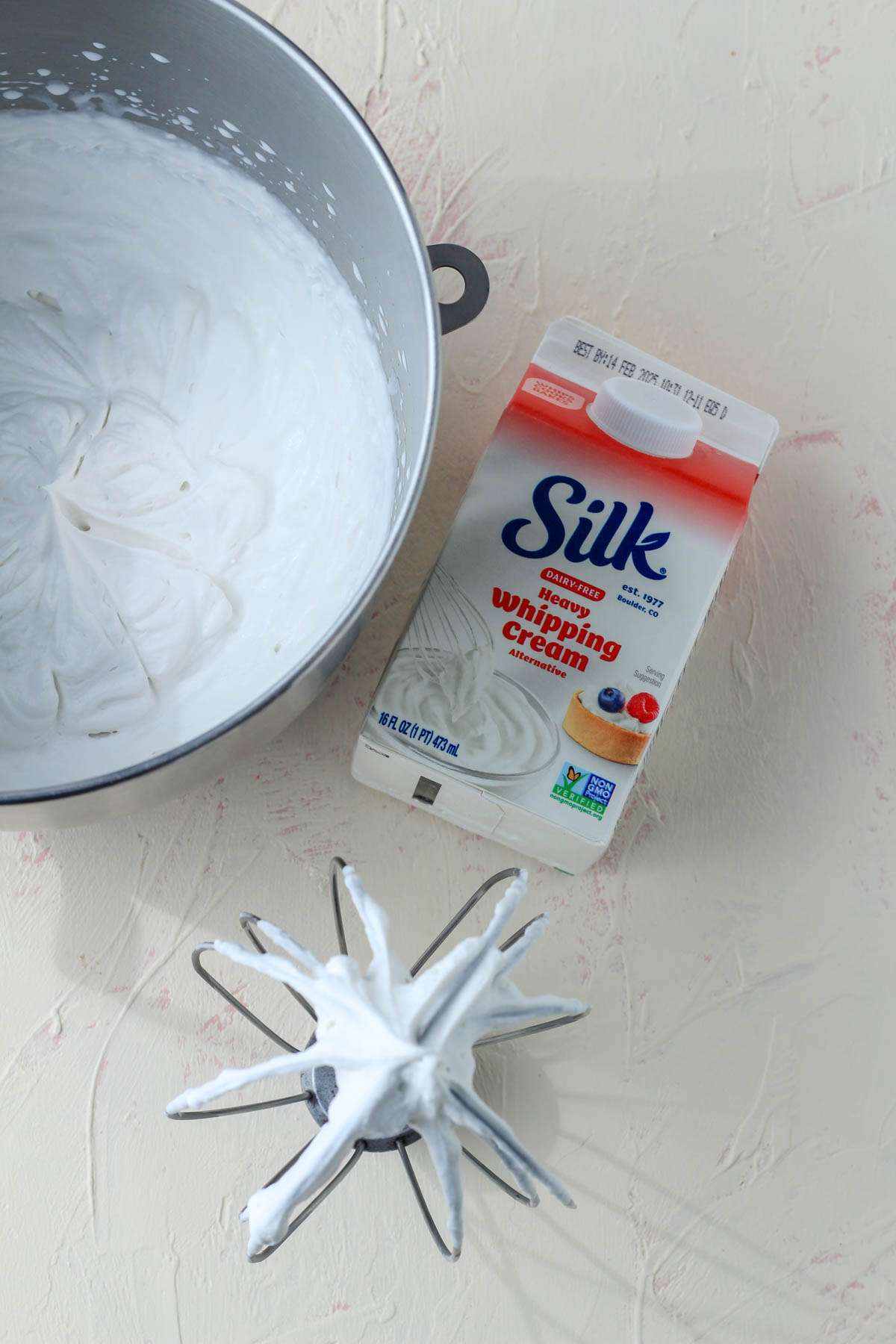 A silver bowl of dairy-free heavy whipped cream with the silk carton to the right and a wire whisk attachment below.