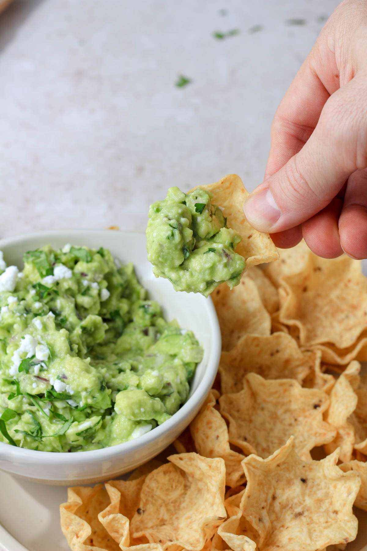 A hand with a scoop of avocado and feta dip over a plate of tortilla scoops.