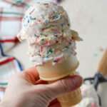 A close up of a cake cone topped with two scoops of vegan no churn cake batter ice cream being held up.