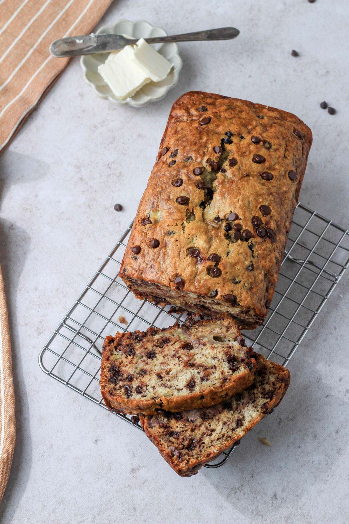 A top down image of a loaf of vegan chocolate chip banana bread on a wire cooling rack with two slices cut off in the front of the loaf and a small dish of vegan butter in the back left.
