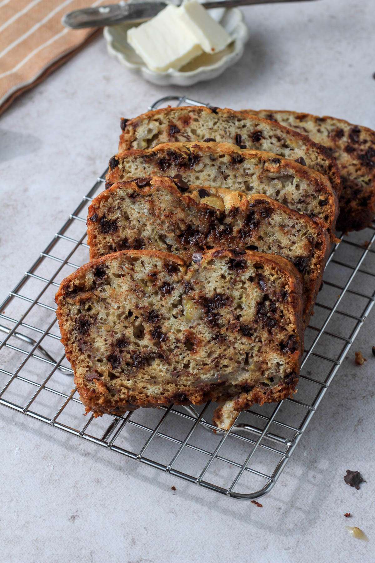 A silver cooling rack with five slices of vegan chocolate chip banana bread laid horizontally with a small dish of vegan butter in the back left.