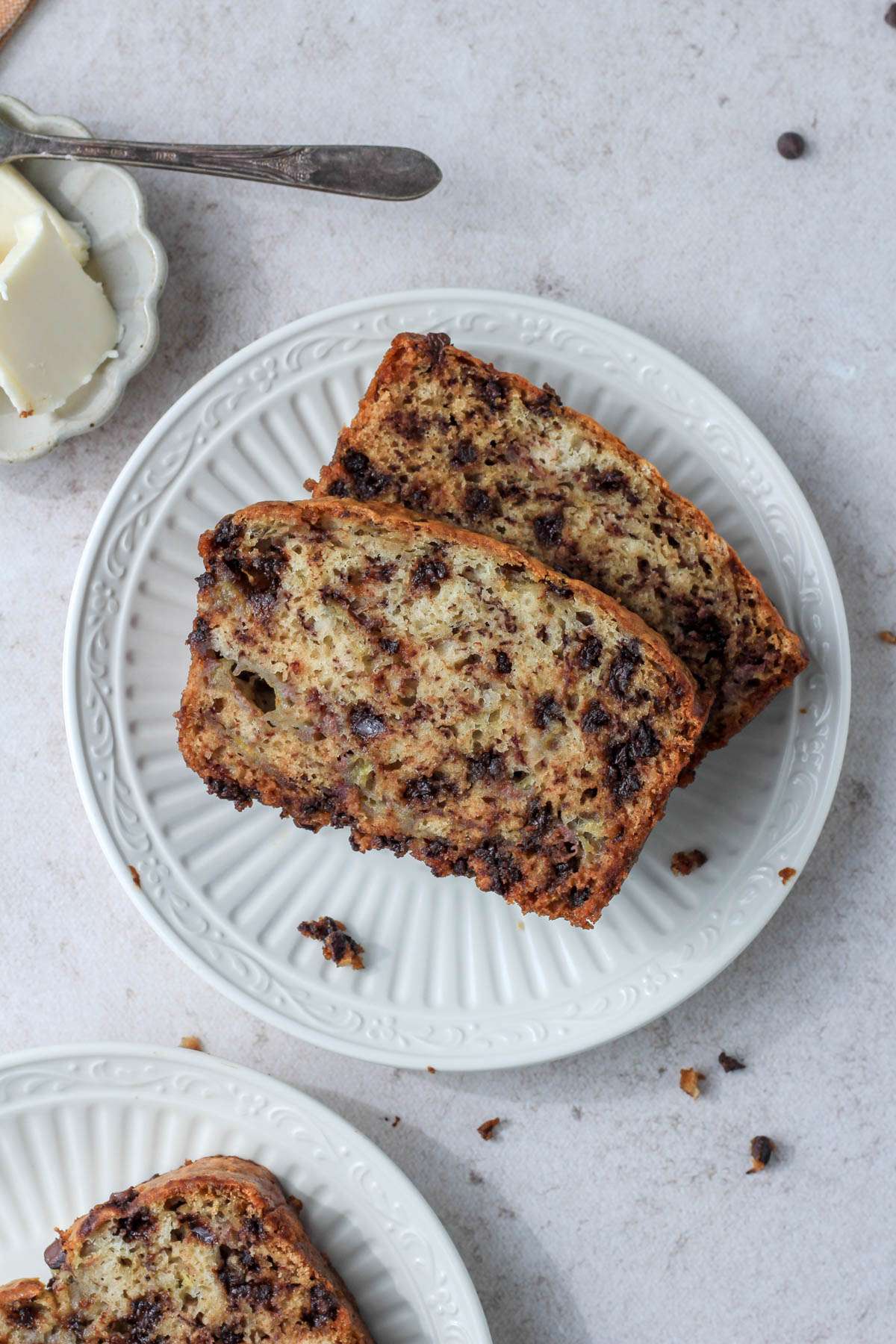 Two white plates with slices of vegan chocolate chip banana bread on a tan counter with a small dish of vegan butter in the top left corner.