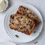 A cropped image of a small white plate topped with two slices of vegan chocolate chip banana bread on a tan counter.
