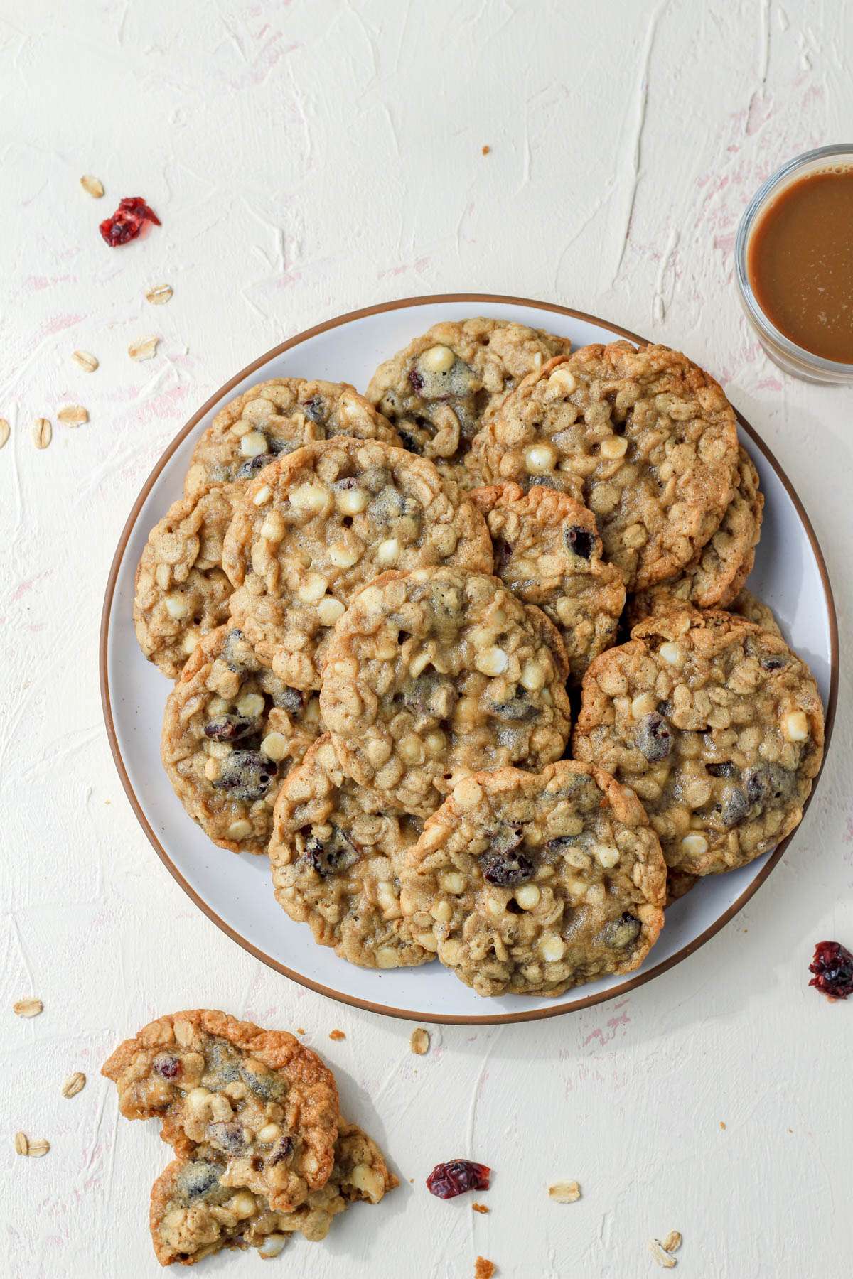 A top down photo of white chocolate chip craisin oatmeal cookies on a cream counter with a broken cookie in the bottom left and a small bowl in the top right.