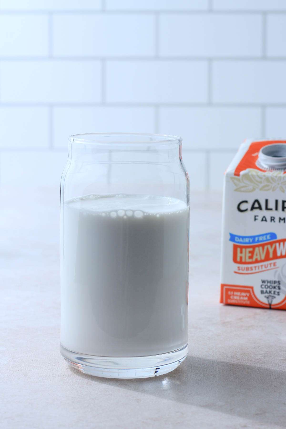 A tall glass of Califia Farms Heavy whip substitute with the orange and white carton in the back right.