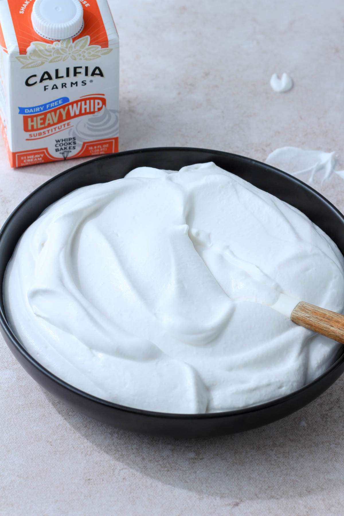 A black bowl of whipped cream with a white and wooden spatula in front of an orange and white carton of Califia Farms Heavy whip in the back left.
