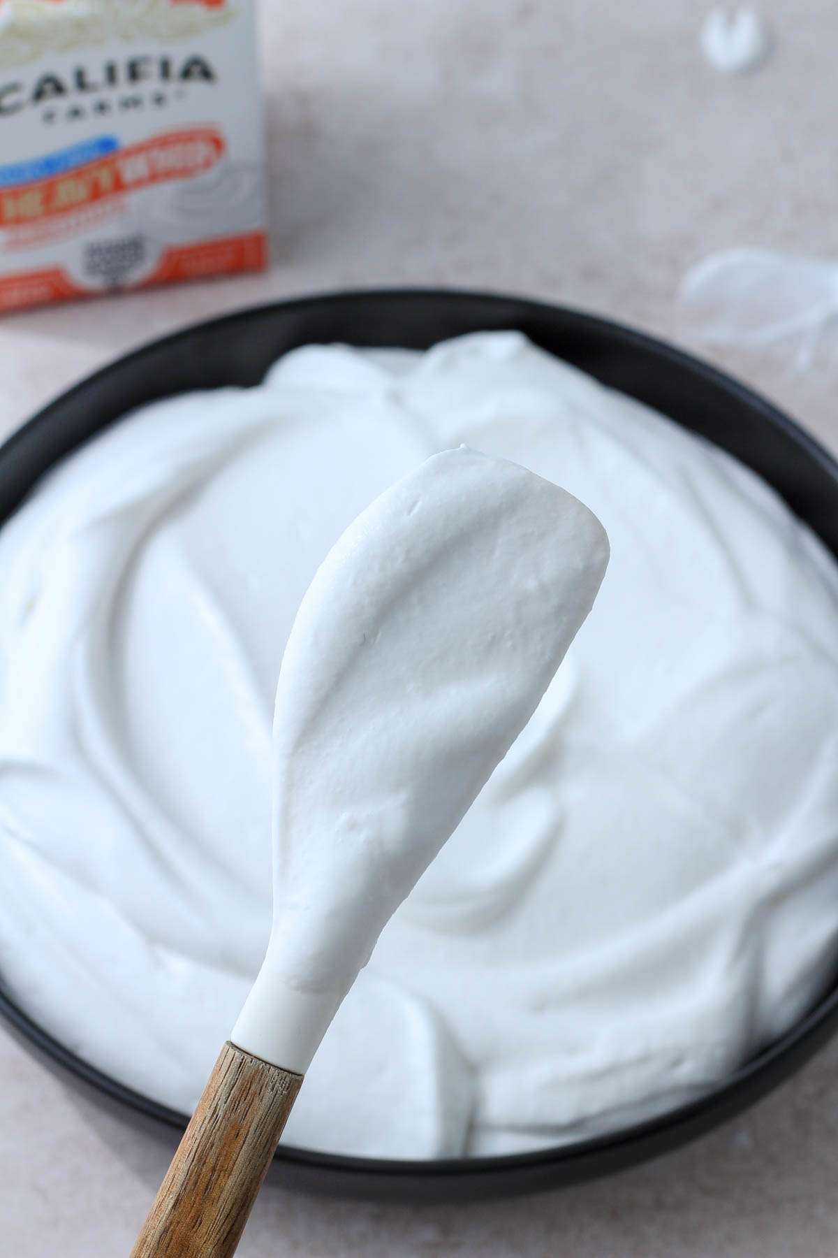 A white spatula topped with heavy whip substitute that has been made into whipped cream over a black bowl of whipped cream with a carton in the back left.