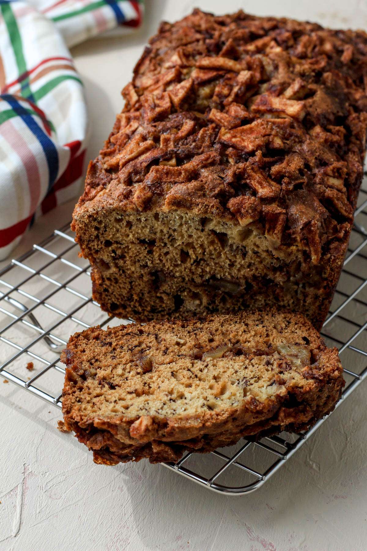 A silver cooling rack with a loaf of cinnamon apple banana bread with a slice cut off in the front.