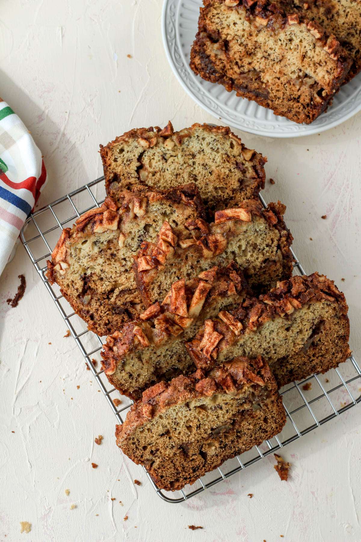 A cream counter with a cooling rack of sliced banana apple bread with a small white plate with a slice of banana bread in the top right.