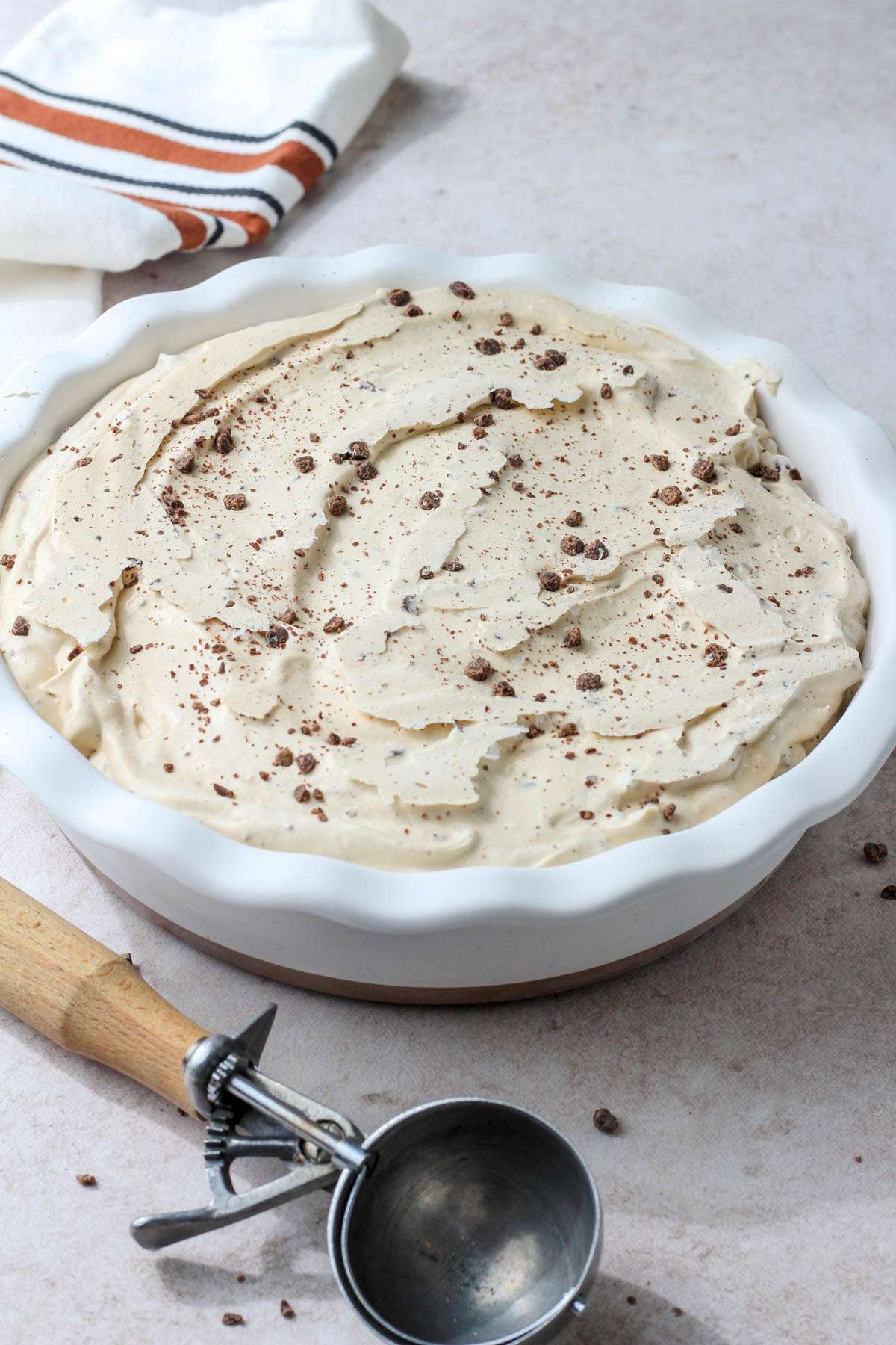 From the side, a white pie pan filled with dairy-free coffee chip ice cream on a tan counter with an ice cream scoop.