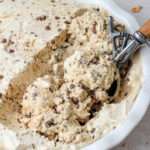 A close up of a white pan filled with coffee chip no churn ice cream scooped with a silver and wooden ice cream scooper.