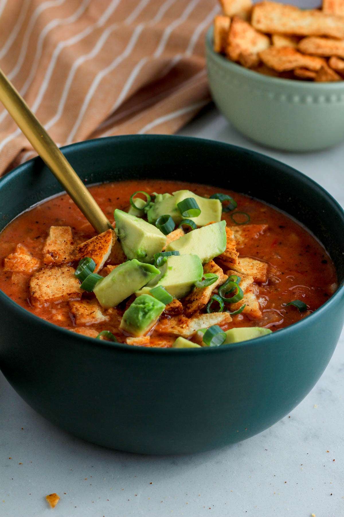 From the side, a dark green bowl with creamy chicken tortilla soup topped with tortilla chips, avocado chunks, and a small bowl of tortilla chips in the back right.