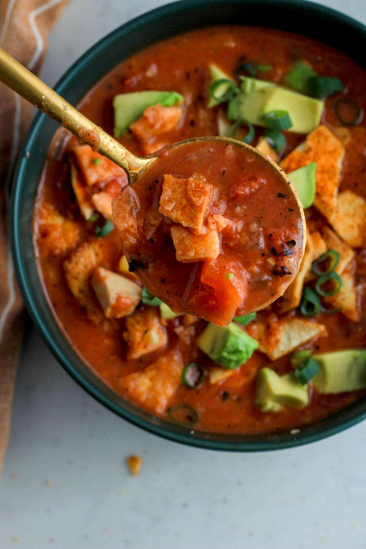 A gold spoon with a scoop of chicken tortilla soup hovering over a dark green bowl of chicken tortilla soup on a white counter.
