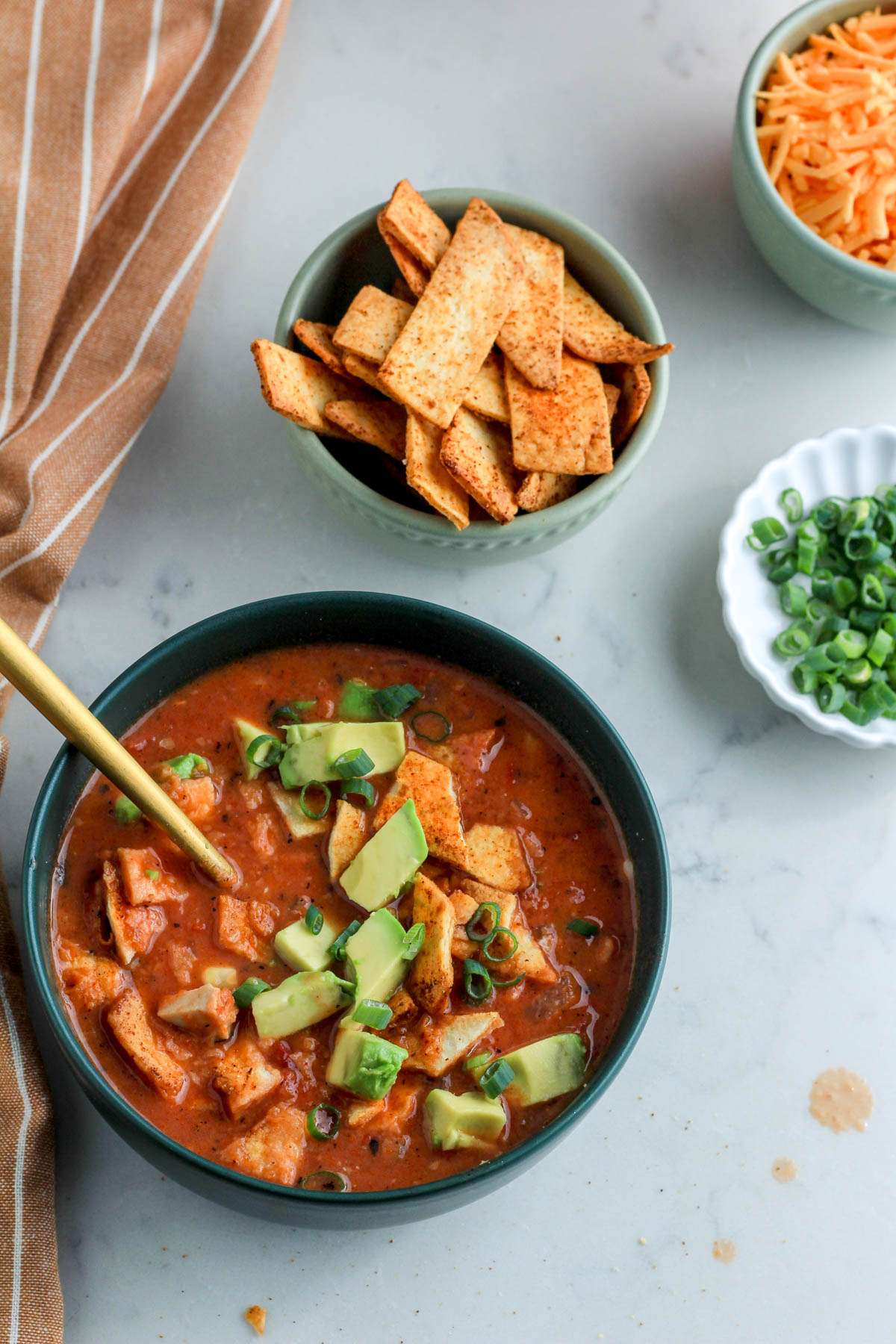 A green bowl of southwest chicken tortilla soup in the bottom left corner with smaller bowls in the top right corner of cheese, tortilla chips, and green onion.