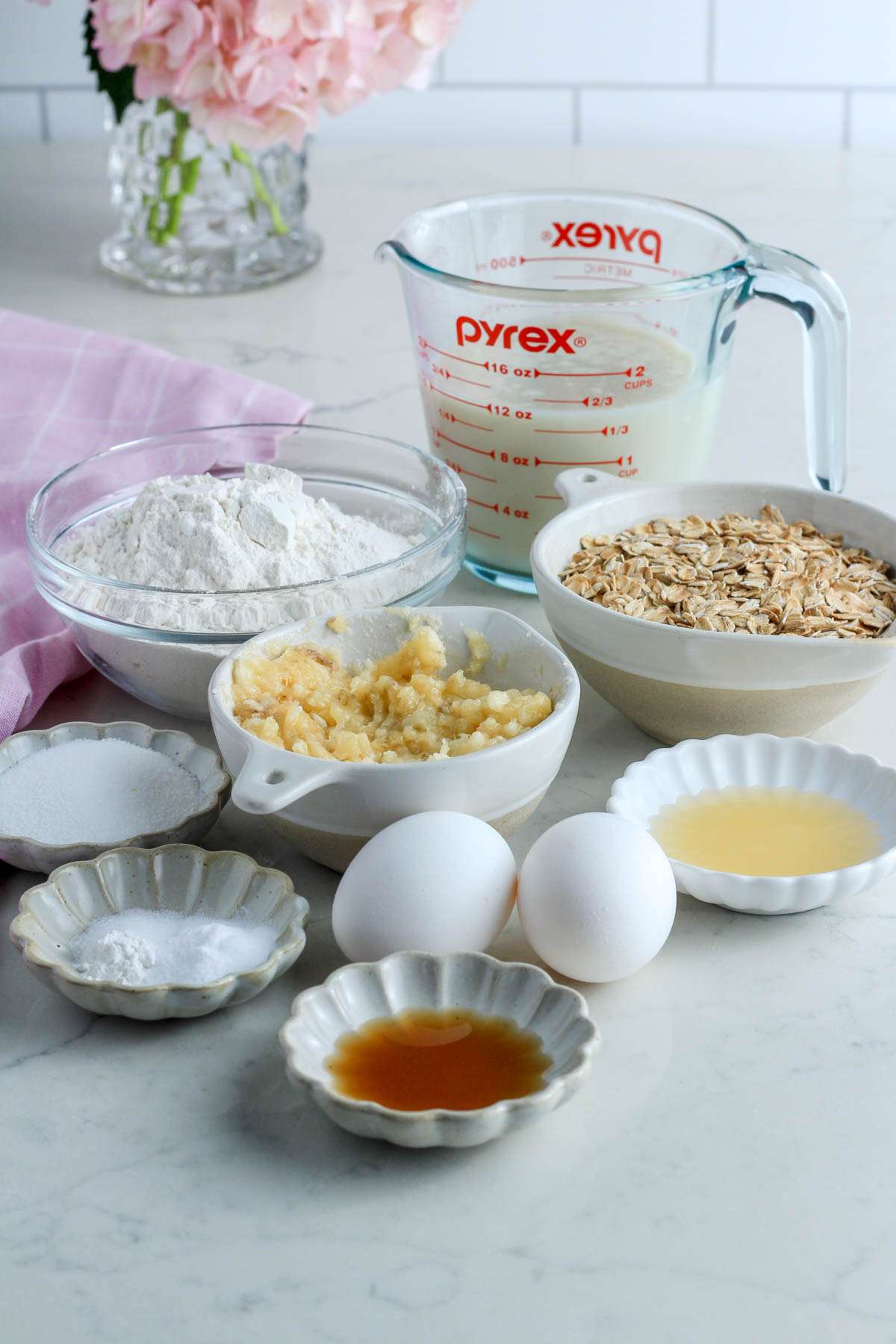 Ingredients for dairy-free oatmeal pancakes on a white counter with a pink towel to the left.