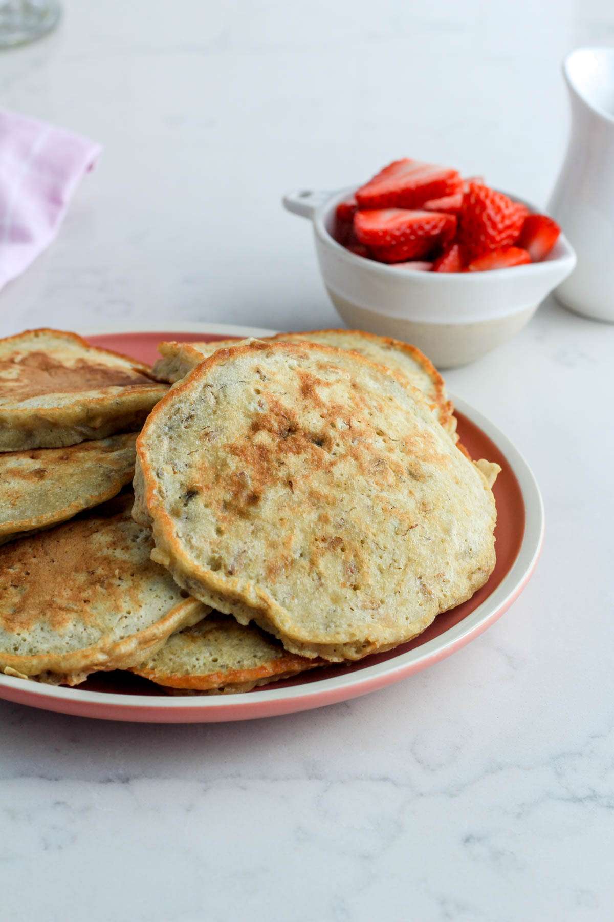 A pink plate with dairy-free oatmeal pancakes on a white counter with a small bowl of sliced strawberries in the back right.