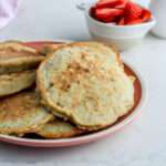 A close up of a pink plate with a pile of dairy-free oatmeal pancakes with a small white bowl of strawberries in the back right.