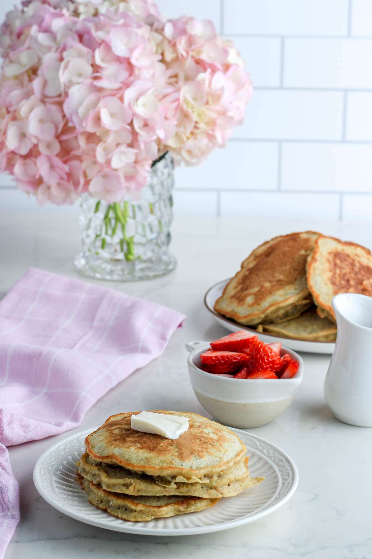 A white plate with three oatmeal pancakes in front of a bowl of strawberries, a jug of syrup, and a plate with a pile of pancakes.