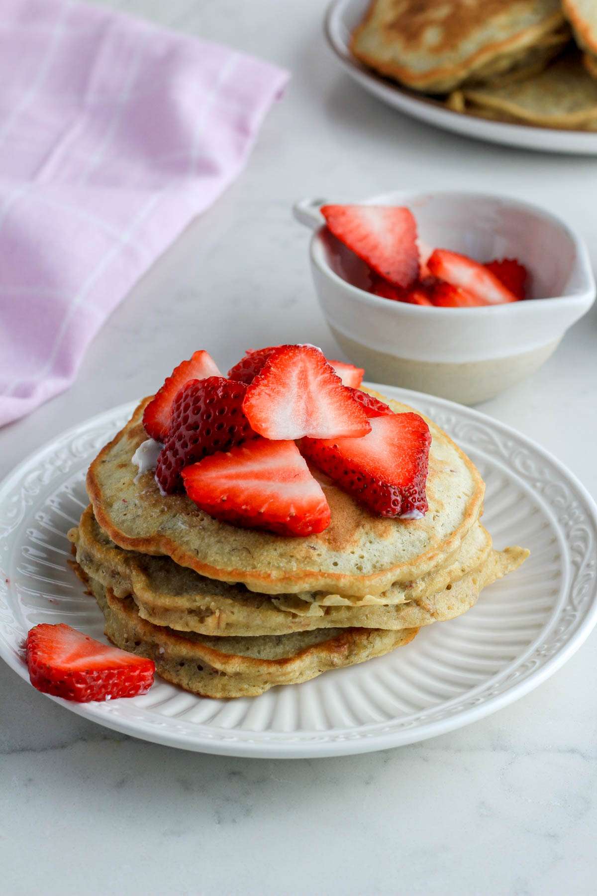A white plate with oatmeal pancakes topped with sliced strawberries in front of a small bowl of sliced strawberries with a white plate of pancakes in the back.