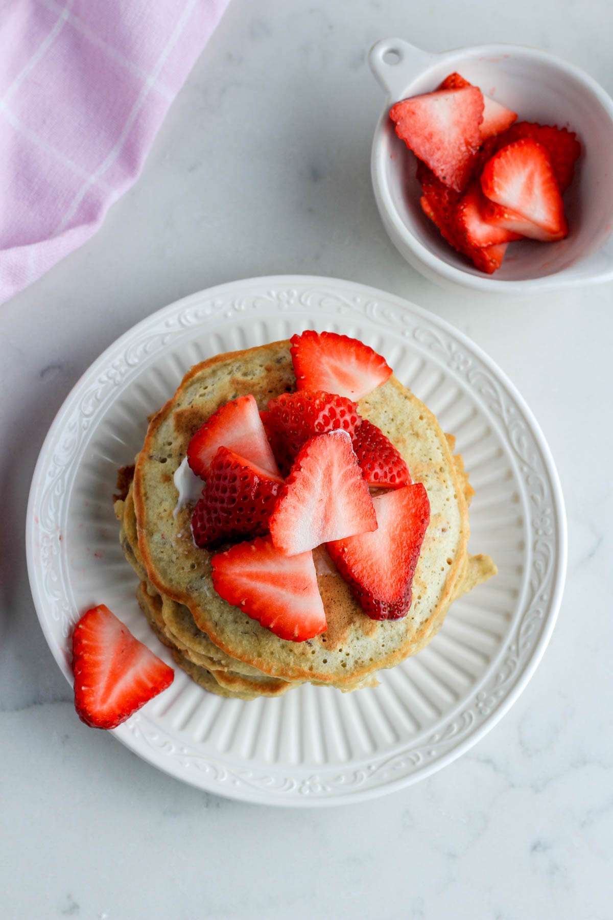 A top down image of a white plate with a stack of oatmeal pancakes topped with sliced strawberries with a small white bowl of strawberries in the top right.