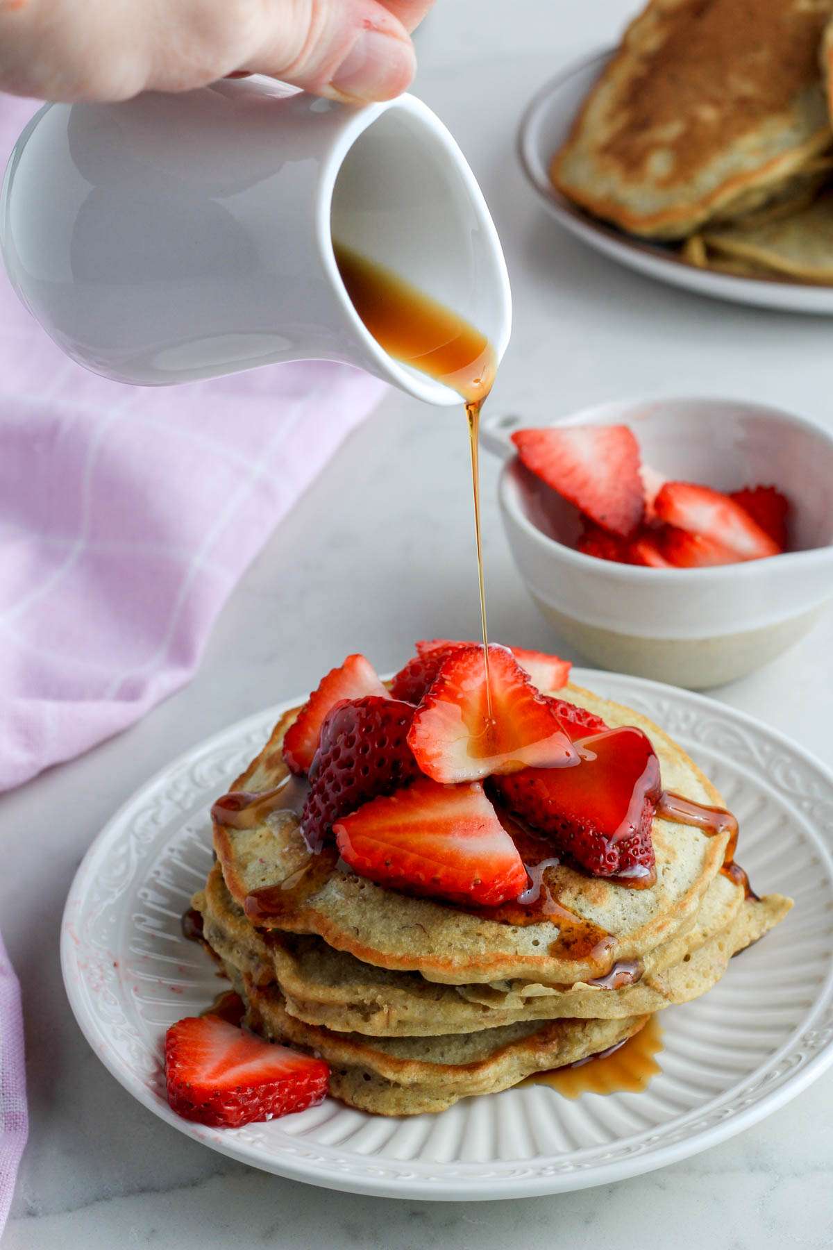 A white plate with three pancakes topped with sliced strawberries with a hand pouring syrup on top of the pancake pile.