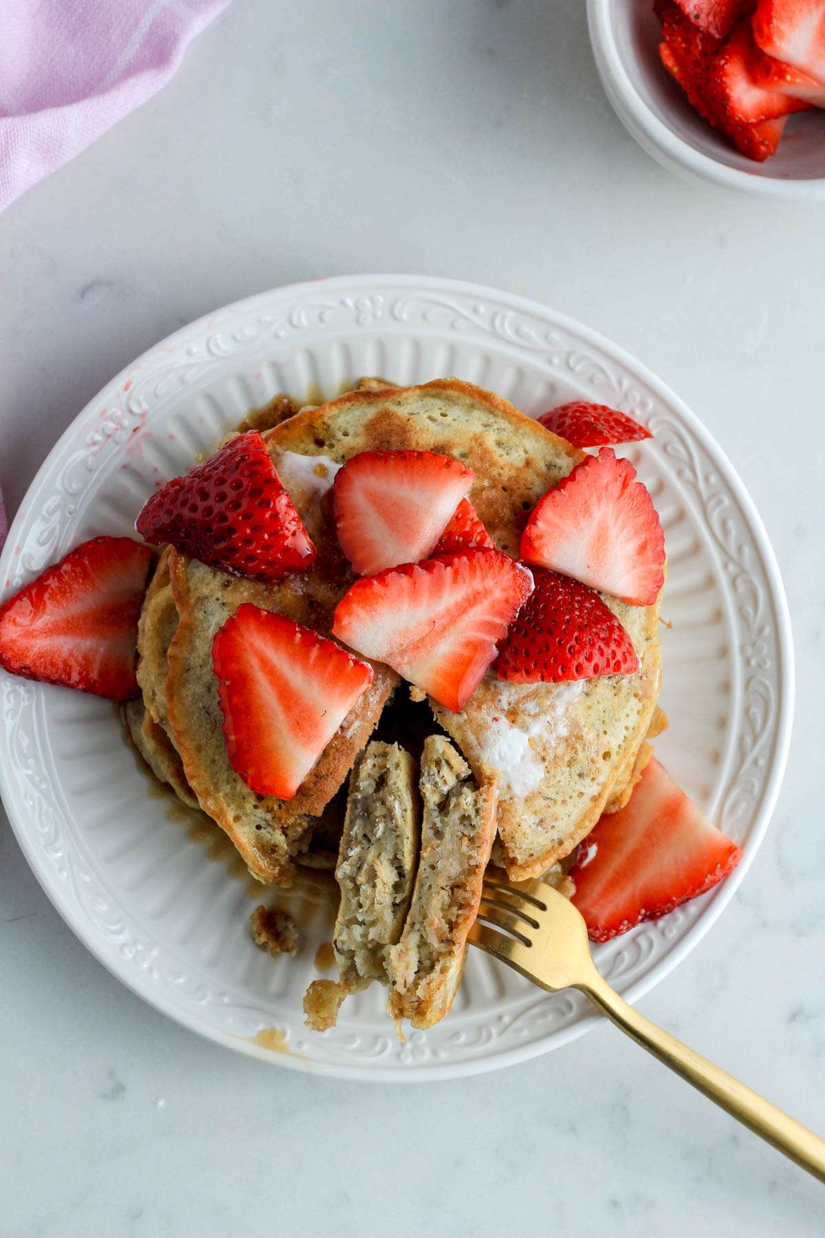 A top down image of a white plate with oatmeal pancakes topped with strawberries and syrup with a wedge cut out with a gold fork.