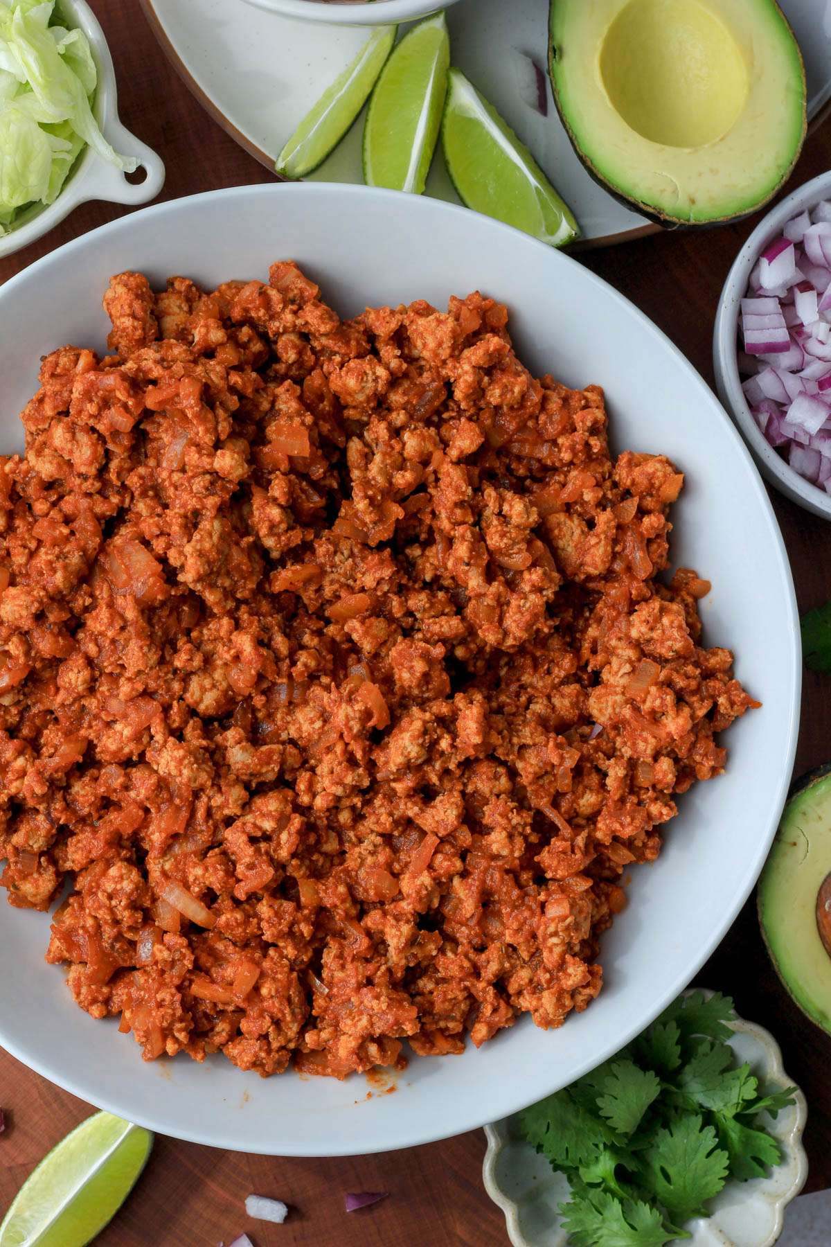 A white bowl of ground turkey taco meat on a wooden counter with taco toppings surrounding the bowl.