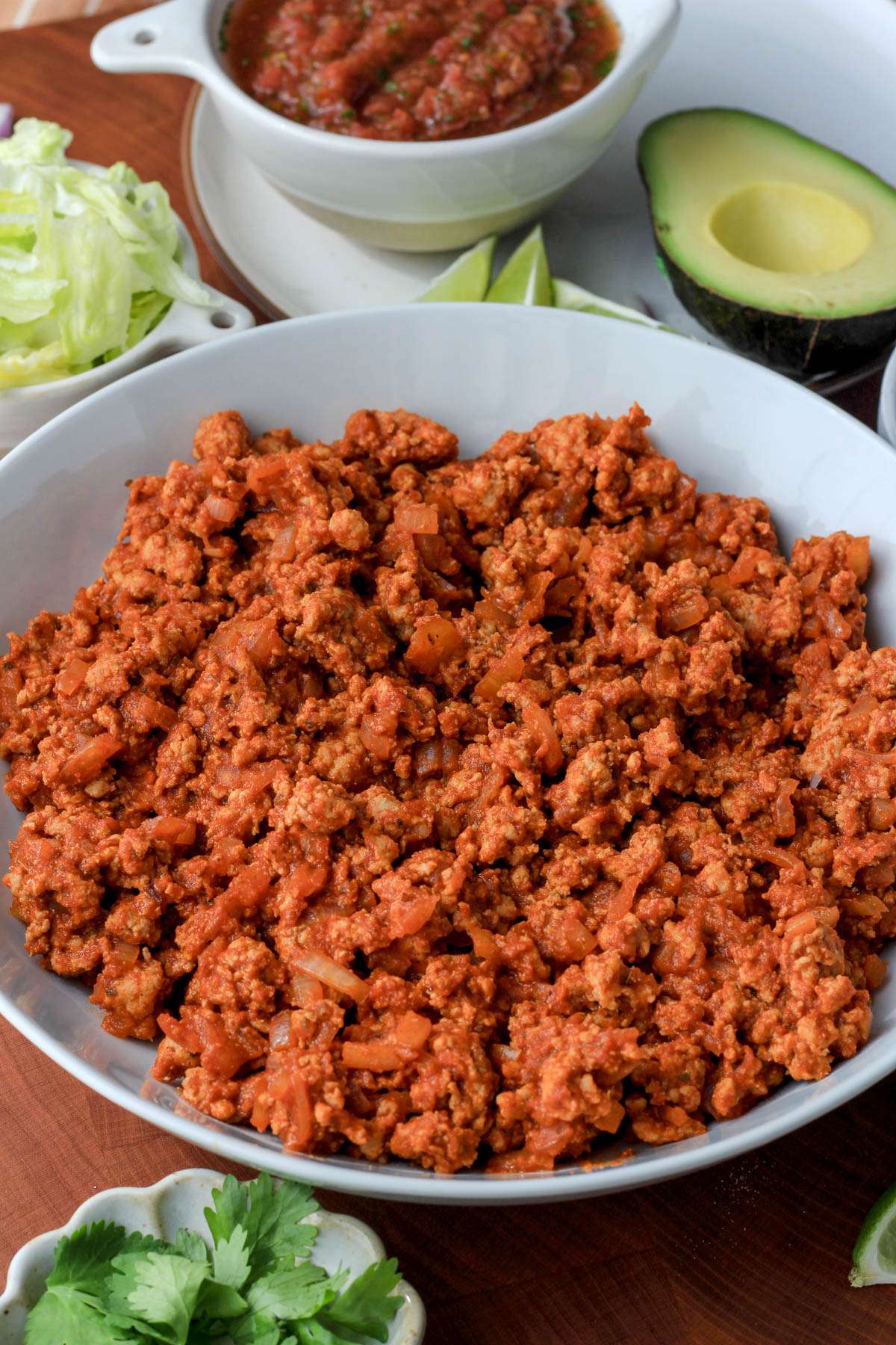 From the side, a white bowl filled with ground turkey taco meat in front of a bowl of salsa, avocado, and shredded lettuce.