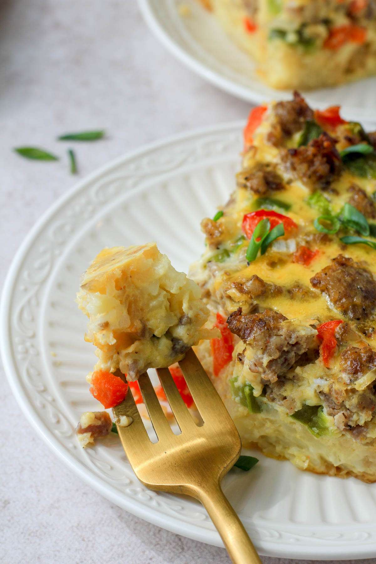 The top of a gold fork with a bite of hashbrown breakfast casserole on a white plate in front of a slice of casserole.