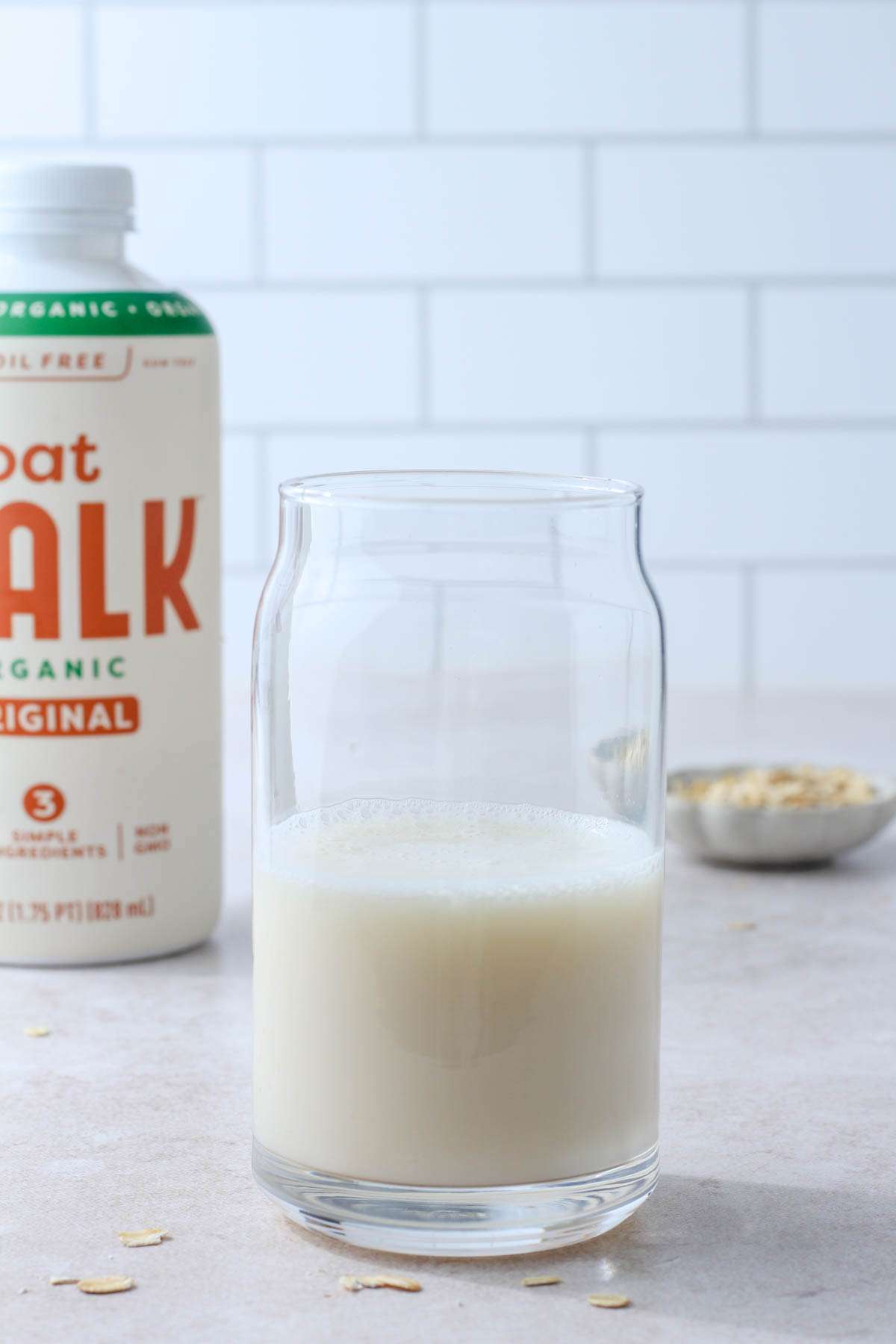 A glass of oat malk in the middle with a bottle of oat malk to the left and a pinch bowl of oats to the right.