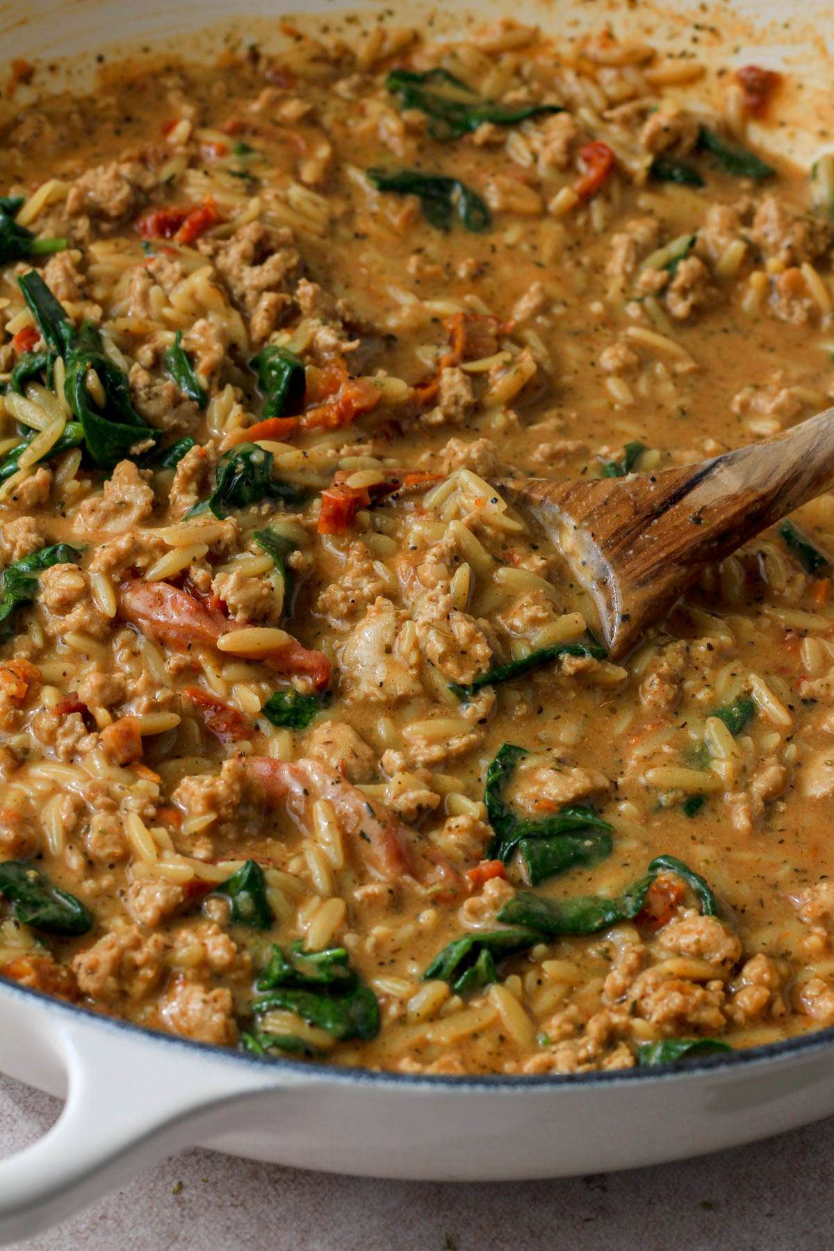A close up of the dairy-free marry me ground chicken and orzo in a white skillet with a wooden spoon scooping the pasta.