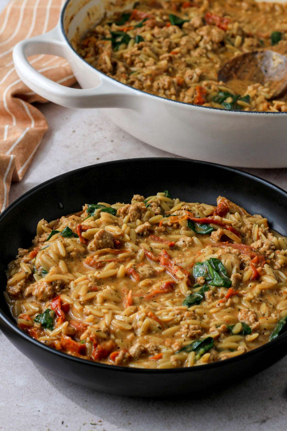 From the side, a black bowl of marry me ground chicken and orzo in front of a white skillet with the rest of the ground chicken and orzo pasta dish.