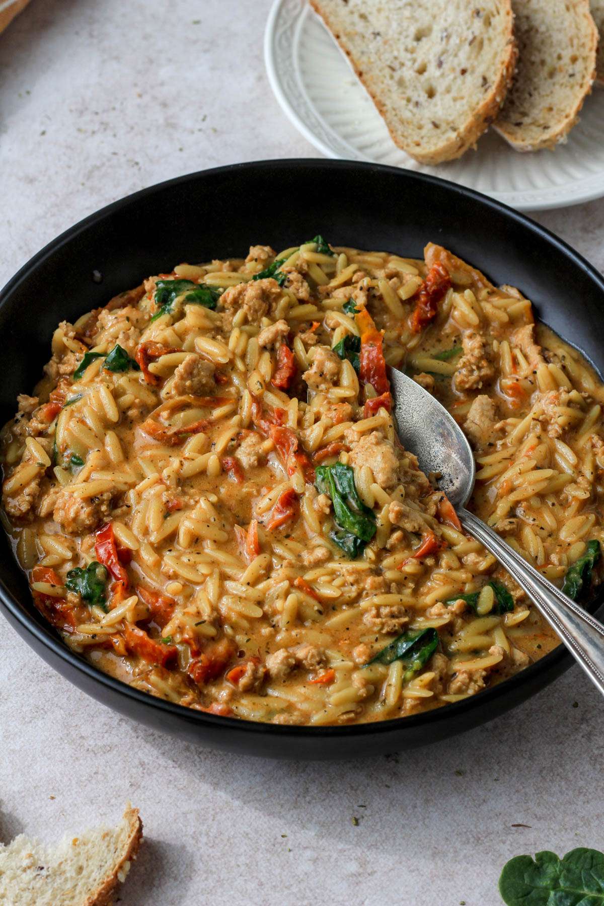 A black bowl with a silver spoon scooping dairy-free marry me ground chicken and orzo in front of a small white plate of bread in the top right.