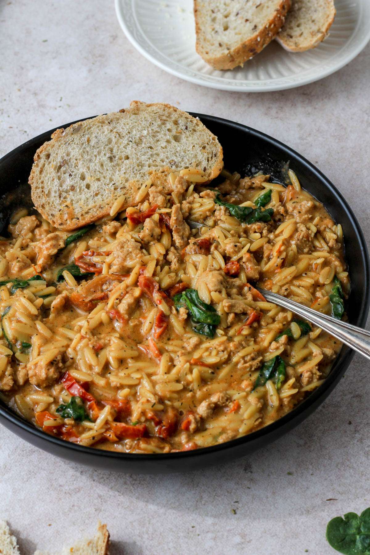 A black bowl of marry me ground chicken and orzo with a silver spoon and a slice of bread in the bowl in front of a small white plate of bread.