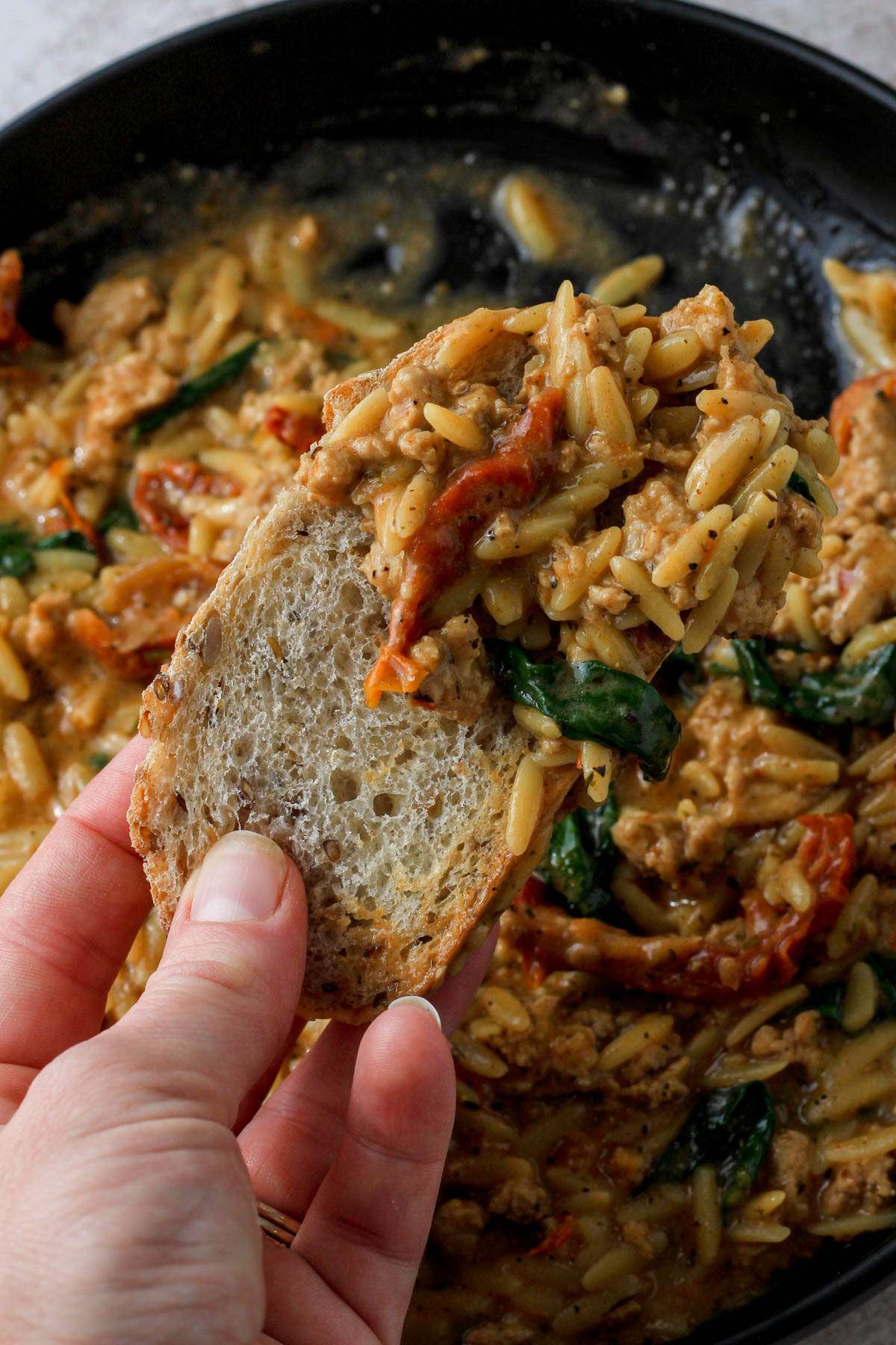 A hand scooping up some of the dairy-free marry me ground chicken and orzo on a piece of bread overt the black bowl.