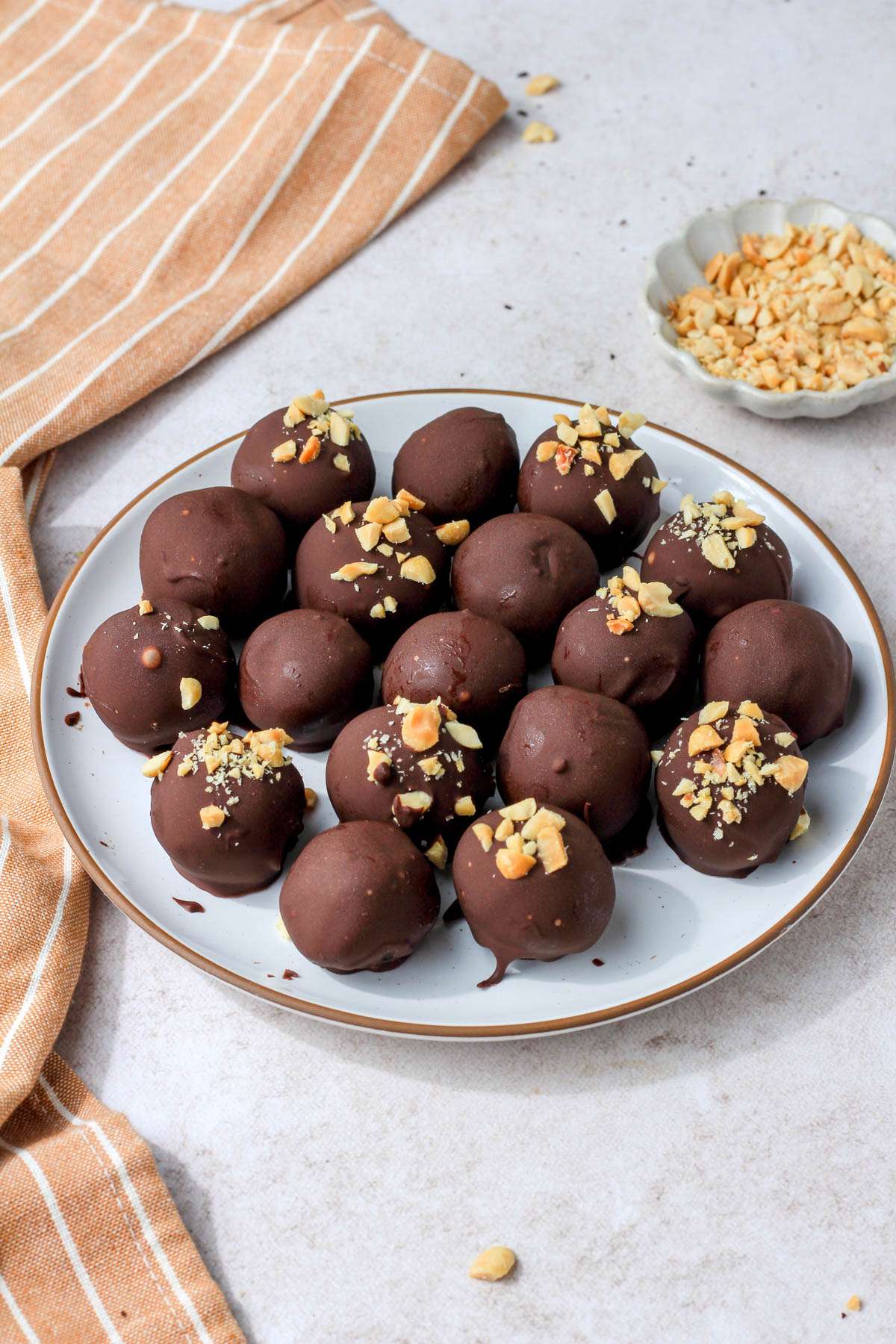 A white plate with a brown rim topped with peanut butter oreo truffles on a tan counter with a small dish of crushed salted peanuts in the back right.