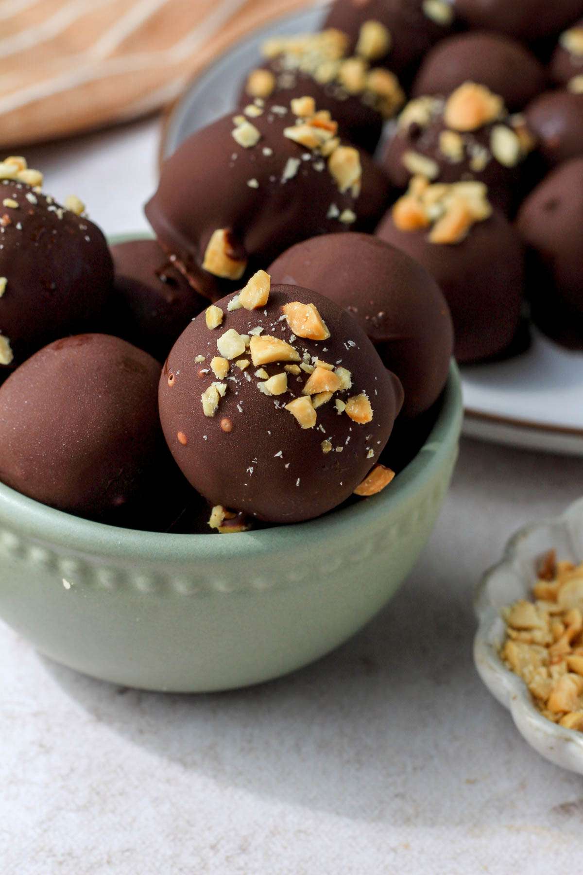 A small green bowl topped with peanut butter oreo truffles in front of a white plate with truffles on a tan counter.