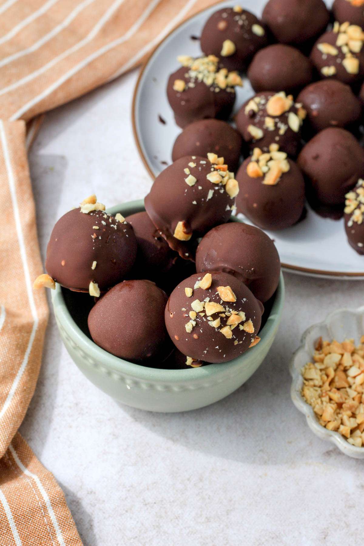 A top down image of a small bowl of peanut butter chocolate oreo truffled in a small green bowl with a white plate of truffles in the back right and a small dish of crushed peanuts to the right.