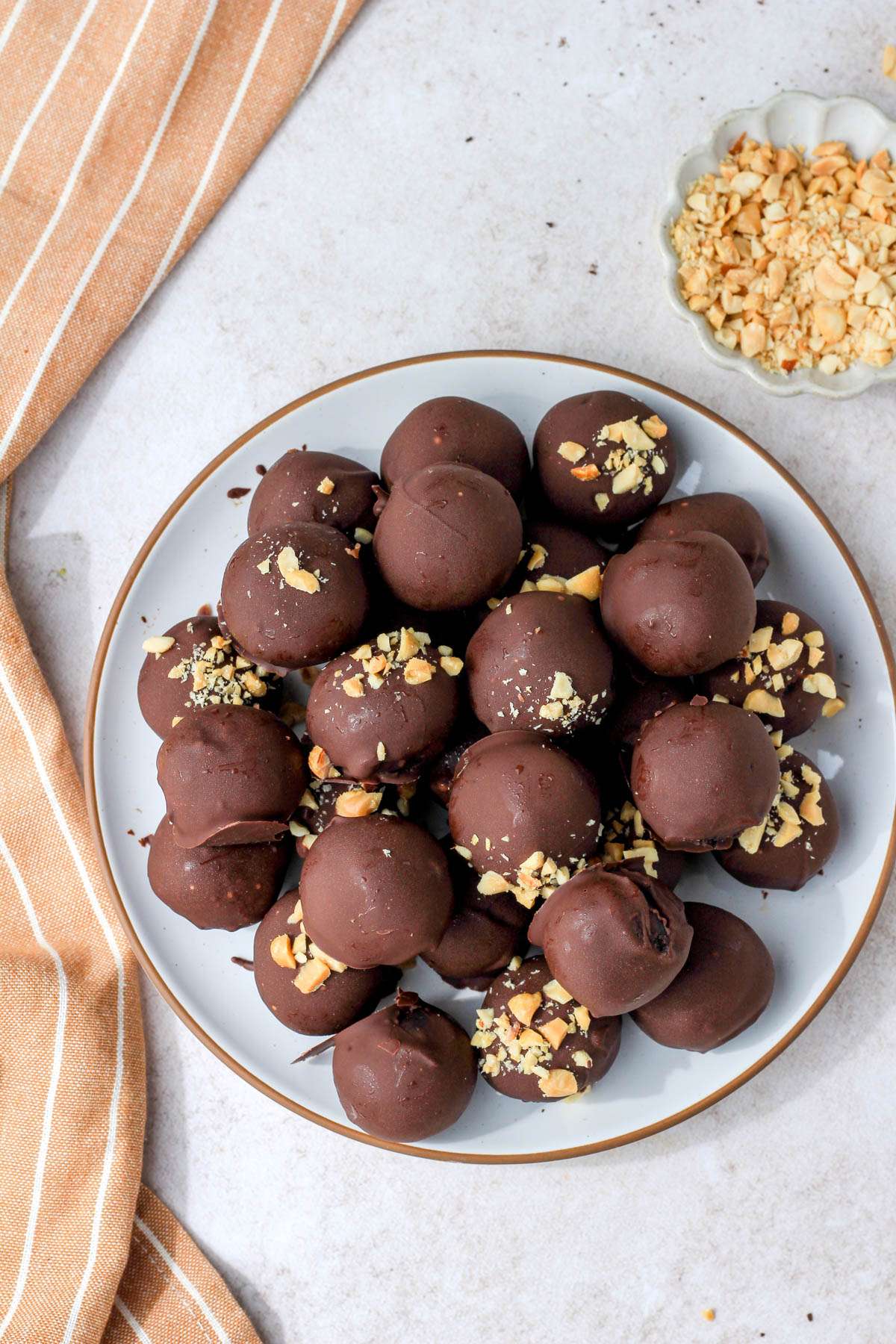 A top down image of a white plate with a brown rim topped with vegan peanut butter oreo truffles on a tan counter with a small dish of crushed salted peanuts in the top right.