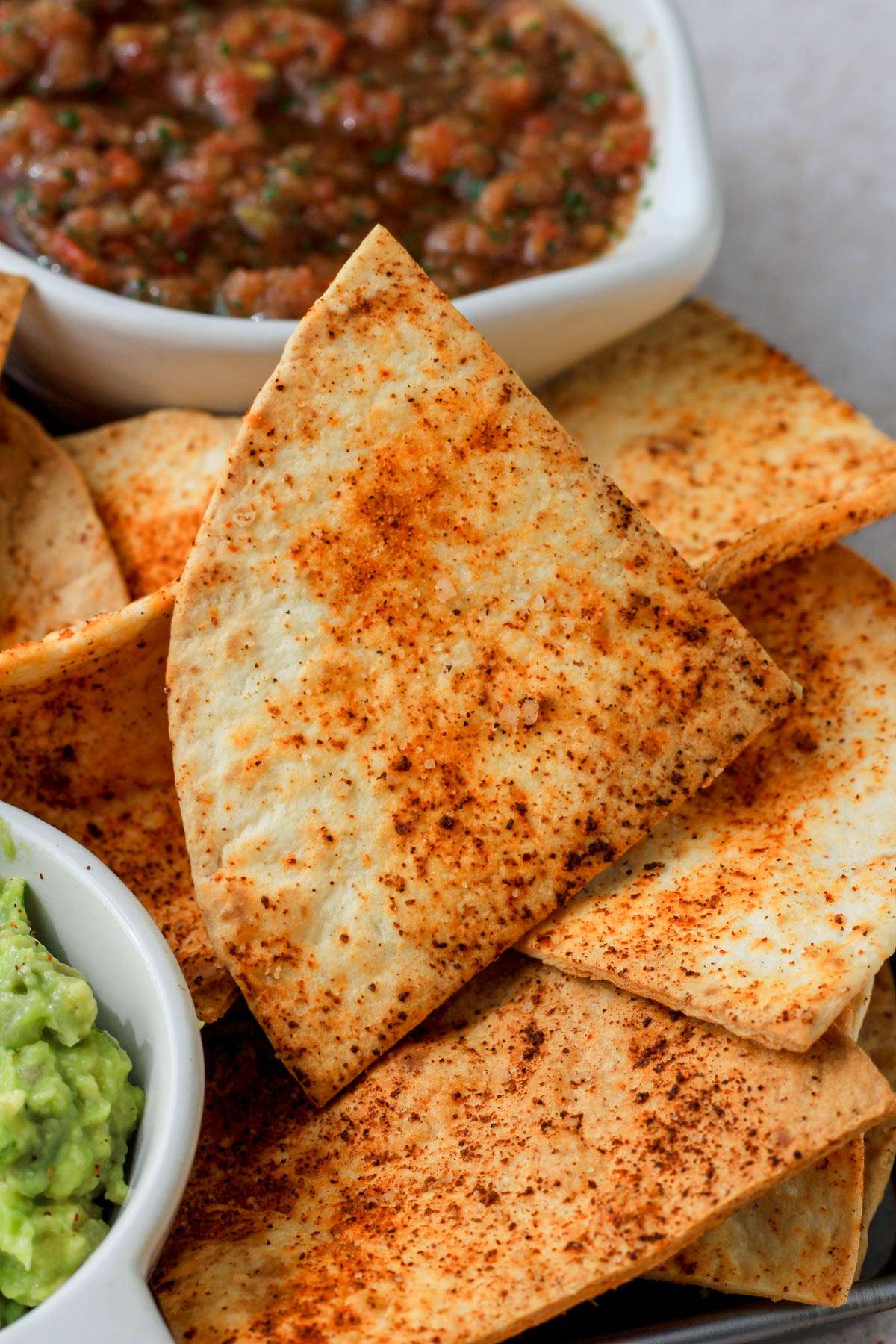 A close up of a seasoned flour tortilla chip on a pile of tortilla chips with a bit of guacamole to the bottom left and some salsa in the top right.