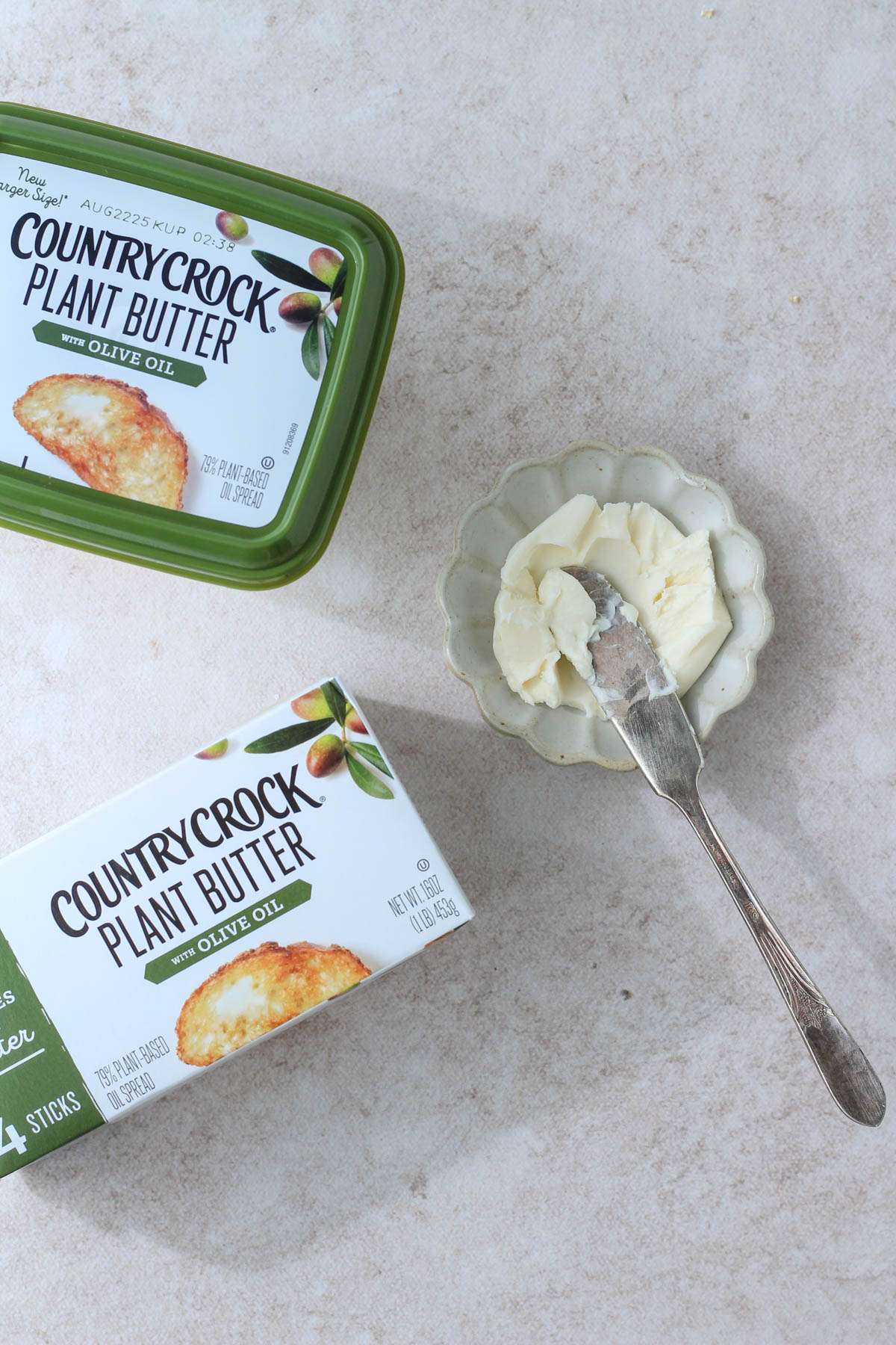 A top down image of two different types of country crock plant based butter on a tan counter with a small dish of butter and a butter knife to the right.